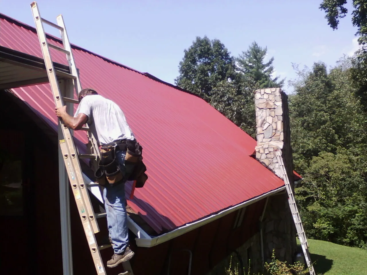Roofer on a ladder beside a red metal roof and stone chimney — Soffit & Fascia Repair by Roofing Brothers in Minocqua